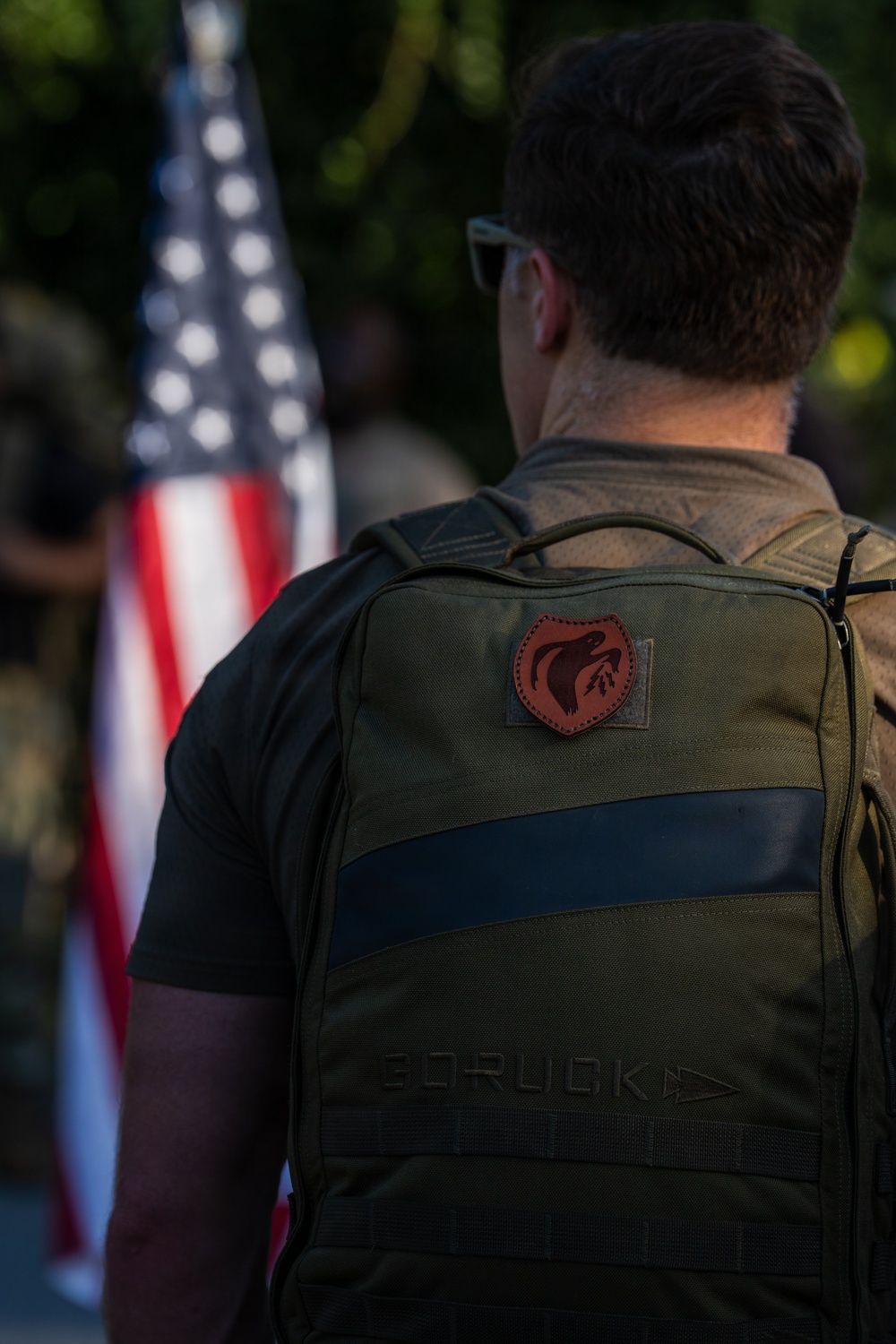 A man is photographed from behind wearing a dark olive/green GORUCK brand backpack with a red shield-shaped patch attached via velcro. An American flag is visible in the blurred background, suggesting a patriotic or military-related event. The image is sharply focused on the backpack and subject, with the background intentionally bokeh.