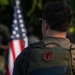 A man is photographed from behind wearing a dark olive/green GORUCK brand backpack with a red shield-shaped patch attached via velcro. An American flag is visible in the blurred background, suggesting a patriotic or military-related event. The image is sharply focused on the backpack and subject, with the background intentionally bokeh.