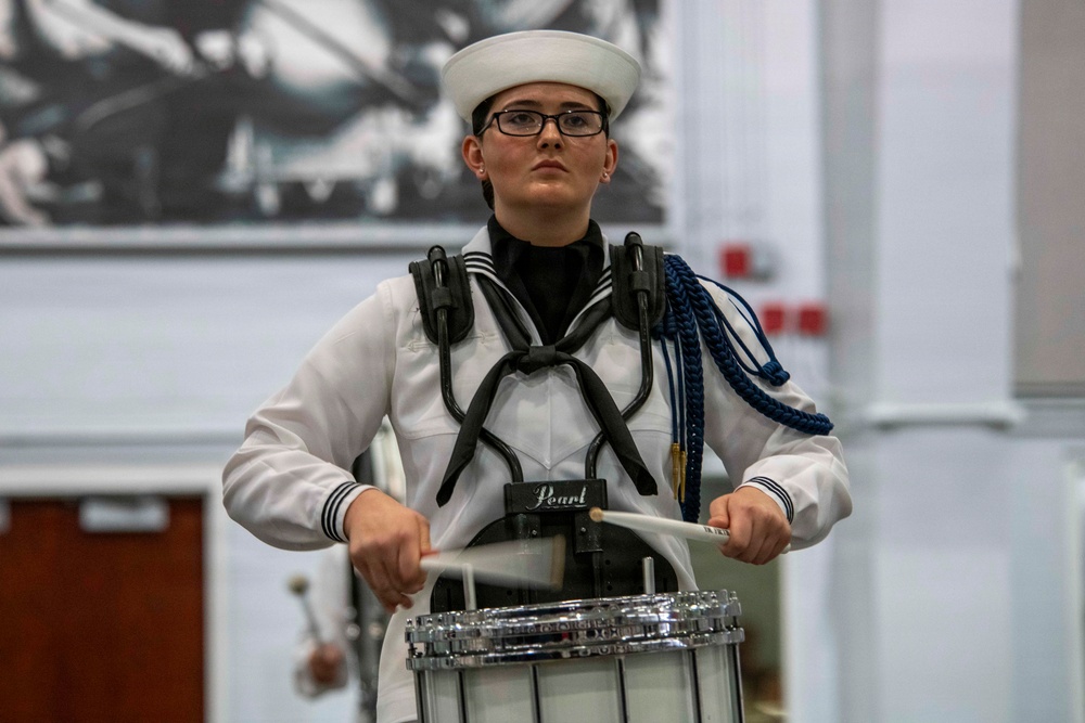 A young person in a white naval-style marching band uniform is playing a Pearl snare drum. They are wearing a white sailor cap, glasses, and have blue decorative cords (aiguillettes) on their uniform. The performer appears focused and serious during what appears to be an indoor marching band performance or competition.