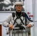 A young person in a white naval-style marching band uniform is playing a Pearl snare drum. They are wearing a white sailor cap, glasses, and have blue decorative cords (aiguillettes) on their uniform. The performer appears focused and serious during what appears to be an indoor marching band performance or competition.