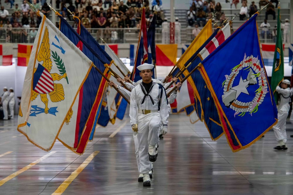 A U.S. Navy color guard ceremony is taking place inside a large indoor arena or gymnasium. Sailors in white dress uniforms are marching in formation while carrying numerous flags representing U.S. states, territories, and military branches. A large audience is visible in the bleachers in the background.