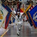 A U.S. Navy color guard ceremony is taking place inside a large indoor arena or gymnasium. Sailors in white dress uniforms are marching in formation while carrying numerous flags representing U.S. states, territories, and military branches. A large audience is visible in the bleachers in the background.