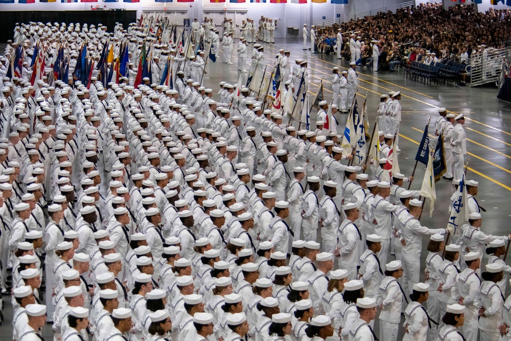 A large-scale U.S. Navy recruit graduation ceremony (Pass in Review) taking place in an indoor arena, likely at Recruit Training Command (RTC) Great Lakes, Illinois. Hundreds of sailors in white dress uniforms are arranged in precise formation columns across the drill floor. Flag bearers carrying unit guidons and national/state flags are visible, with a large audience of civilians observing from the stands.
