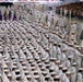 A large-scale U.S. Navy recruit graduation ceremony (Pass in Review) taking place in an indoor arena, likely at Recruit Training Command (RTC) Great Lakes, Illinois. Hundreds of sailors in white dress uniforms are arranged in precise formation columns across the drill floor. Flag bearers carrying unit guidons and national/state flags are visible, with a large audience of civilians observing from the stands.