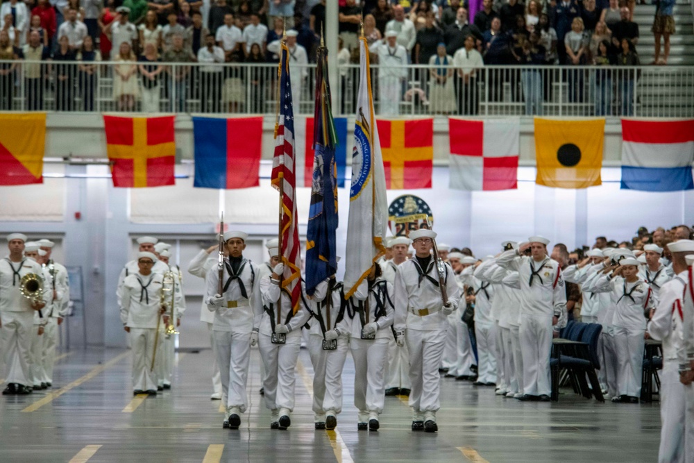 A formal U.S. Navy color guard ceremony is taking place inside a large indoor venue, likely a military training facility or gymnasium. Personnel in dress white uniforms are carrying flags including the American flag and what appears to be a Navy command flag, while surrounding sailors render salutes. A large audience of spectators observes from bleachers in the background.