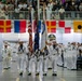 A formal U.S. Navy color guard ceremony is taking place inside a large indoor venue, likely a military training facility or gymnasium. Personnel in dress white uniforms are carrying flags including the American flag and what appears to be a Navy command flag, while surrounding sailors render salutes. A large audience of spectators observes from bleachers in the background.