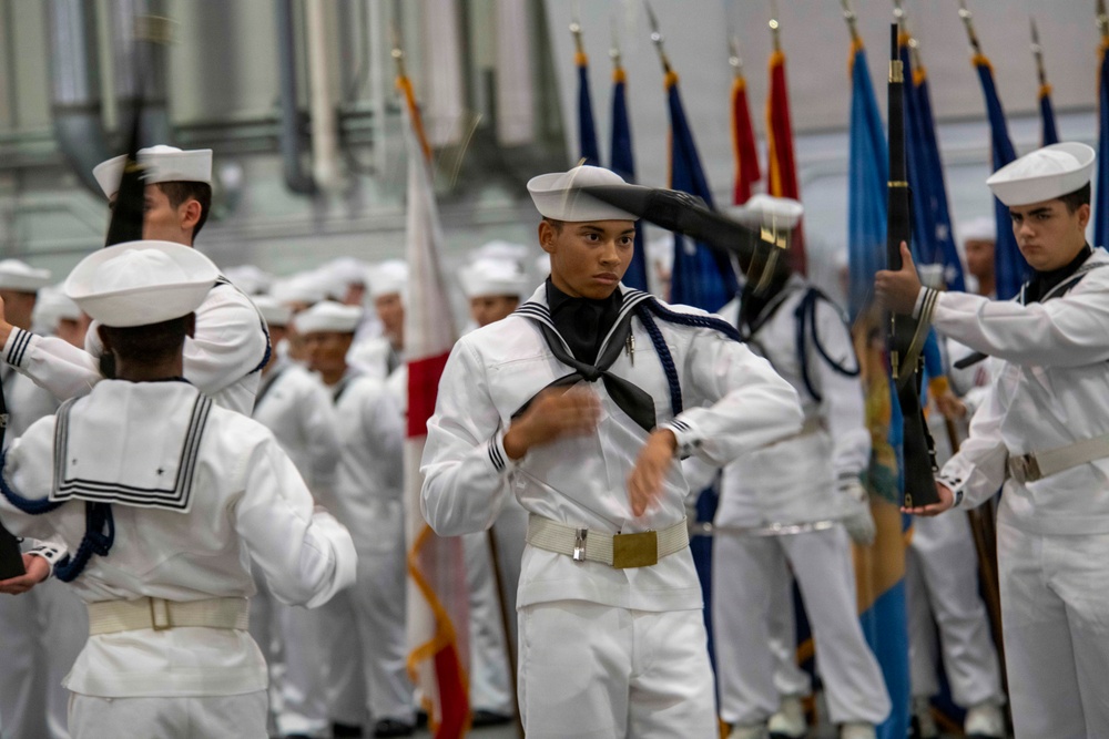 The image shows a group of naval personnel, likely recruits or cadets, dressed in traditional white Navy dress uniforms with white sailor caps and black neckerchiefs. They appear to be participating in a formal drill or graduation ceremony, with a color guard holding multiple flags visible in the background. The central figure is captured mid-motion, suggesting an active drill movement or marching sequence.