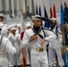 The image shows a group of naval personnel, likely recruits or cadets, dressed in traditional white Navy dress uniforms with white sailor caps and black neckerchiefs. They appear to be participating in a formal drill or graduation ceremony, with a color guard holding multiple flags visible in the background. The central figure is captured mid-motion, suggesting an active drill movement or marching sequence.