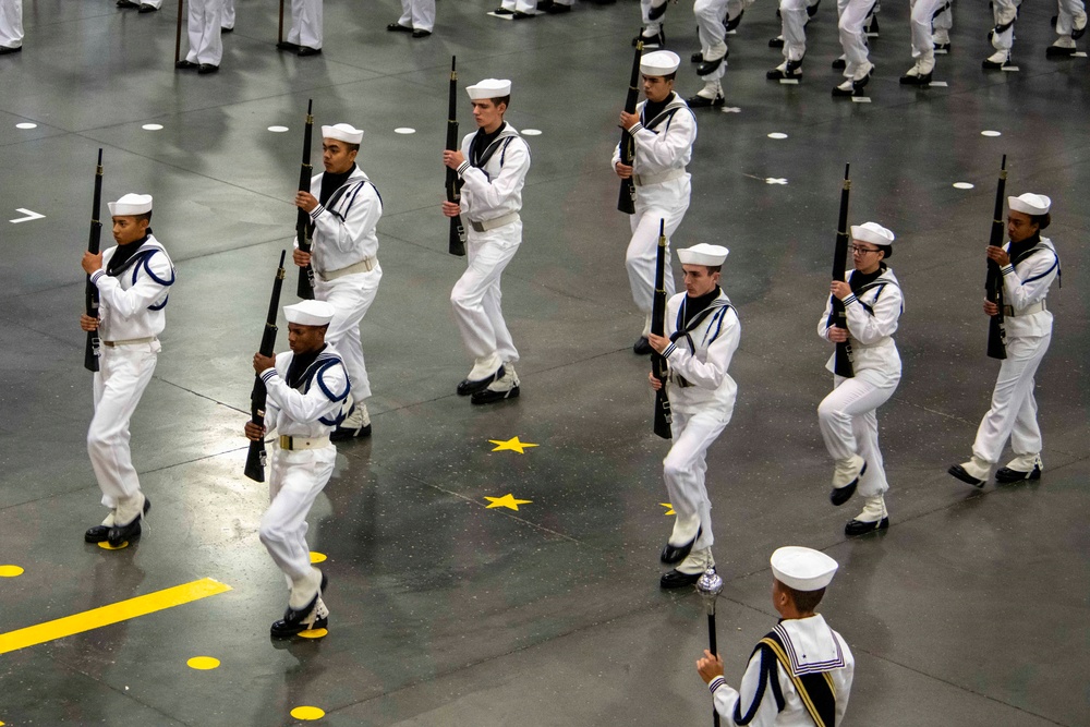 The image shows a large group of naval recruits or sailors in white dress uniforms performing a formal drill ceremony indoors. They are carrying rifles and marching in coordinated formation on a large indoor floor marked with yellow stars and dots. The event appears to be a military graduation or pass-in-review ceremony.
