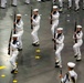 The image shows a large group of naval recruits or sailors in white dress uniforms performing a formal drill ceremony indoors. They are carrying rifles and marching in coordinated formation on a large indoor floor marked with yellow stars and dots. The event appears to be a military graduation or pass-in-review ceremony.