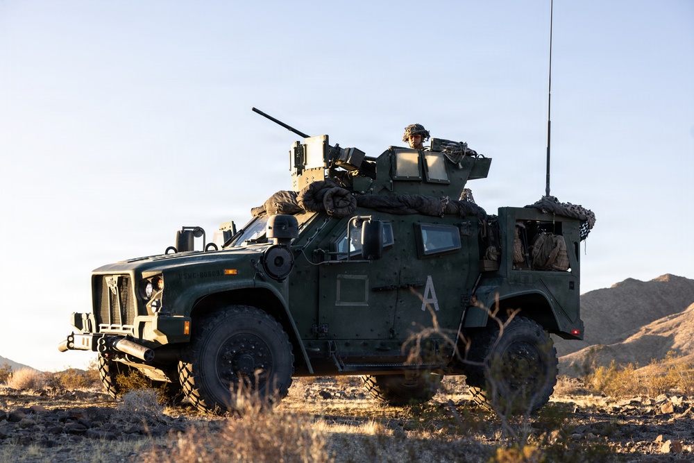 A military tactical vehicle, identified as a Joint Light Tactical Vehicle (JLTV), is positioned in an arid desert terrain during golden hour lighting. At least one soldier is visible manning a weapon system mounted on the vehicle's turret. The vehicle bears the letter 'A' on its door and appears to be on a field exercise or patrol operation.