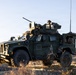 A military tactical vehicle, identified as a Joint Light Tactical Vehicle (JLTV), is positioned in an arid desert terrain during golden hour lighting. At least one soldier is visible manning a weapon system mounted on the vehicle's turret. The vehicle bears the letter 'A' on its door and appears to be on a field exercise or patrol operation.