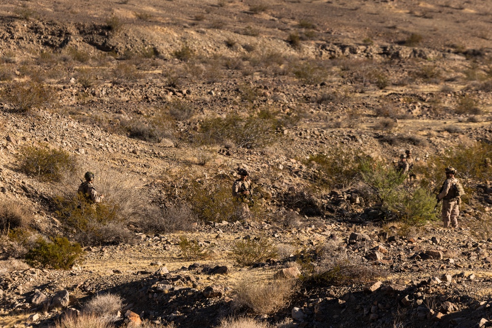 The image shows three military personnel in desert camouflage uniforms conducting a patrol or movement through arid, rocky terrain. The soldiers are spread out in a tactical formation across a dry desert landscape with sparse scrub vegetation. The scene appears to be either a training exercise or an operational patrol in a desert environment.
