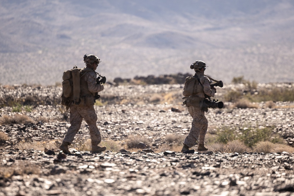 Two military personnel in desert camouflage uniforms are patrolling on foot across a rocky, arid terrain. Both soldiers are fully equipped with tactical gear, helmets, and backpacks, with one visibly carrying a large tube-style weapon system. The background features a vast desert landscape with distant mountains partially obscured by haze.