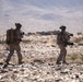 Two military personnel in desert camouflage uniforms are patrolling on foot across a rocky, arid terrain. Both soldiers are fully equipped with tactical gear, helmets, and backpacks, with one visibly carrying a large tube-style weapon system. The background features a vast desert landscape with distant mountains partially obscured by haze.