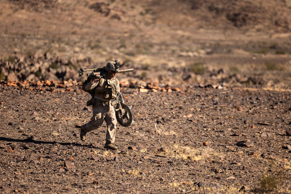 A soldier in desert camouflage uniform is walking across a rocky, arid terrain while carrying what appears to be a shoulder-fired weapon system (likely a Javelin or similar anti-tank missile launcher) on his shoulder and a tripod or equipment stand in his other hand. The environment is a barren desert landscape with scattered rocks and sparse vegetation, suggesting a training exercise or operational movement. The lighting indicates late afternoon or early morning sun casting warm tones across the scene.