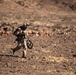 A soldier in desert camouflage uniform is walking across a rocky, arid terrain while carrying what appears to be a shoulder-fired weapon system (likely a Javelin or similar anti-tank missile launcher) on his shoulder and a tripod or equipment stand in his other hand. The environment is a barren desert landscape with scattered rocks and sparse vegetation, suggesting a training exercise or operational movement. The lighting indicates late afternoon or early morning sun casting warm tones across the scene.