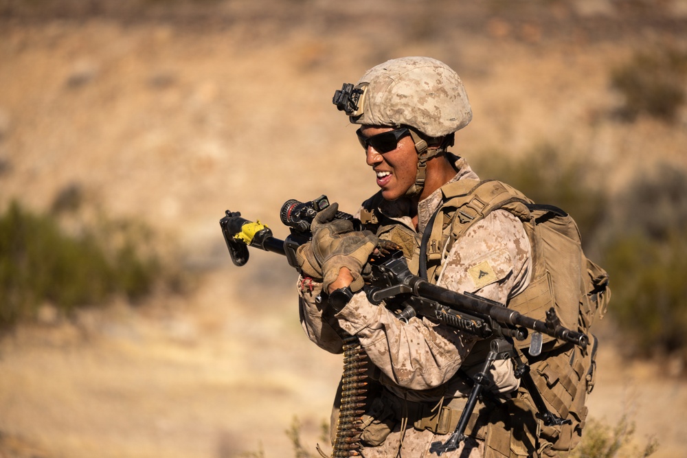 A U.S. Marine in full combat gear is handling a machine gun, likely an M249 SAW or M240B, in an arid desert training environment. The service member is wearing MARPAT desert camouflage, a combat helmet with NVG mount, tactical sunglasses, and a full load-bearing pack. The soldier appears to be inspecting or clearing the weapon, with a visible ammunition belt and a yellow safety flag on the muzzle.