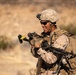 A U.S. Marine in full combat gear is handling a machine gun, likely an M249 SAW or M240B, in an arid desert training environment. The service member is wearing MARPAT desert camouflage, a combat helmet with NVG mount, tactical sunglasses, and a full load-bearing pack. The soldier appears to be inspecting or clearing the weapon, with a visible ammunition belt and a yellow safety flag on the muzzle.