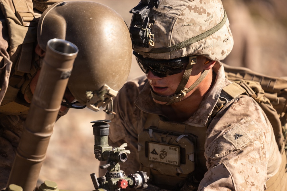 A U.S. Marine in full combat gear is operating what appears to be a mortar system in an arid, desert-like environment. The Marine is wearing MARPAT desert camouflage and is focused on adjusting or sighting the weapon system. A second partially visible service member is present in the background.