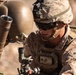 A U.S. Marine in full combat gear is operating what appears to be a mortar system in an arid, desert-like environment. The Marine is wearing MARPAT desert camouflage and is focused on adjusting or sighting the weapon system. A second partially visible service member is present in the background.