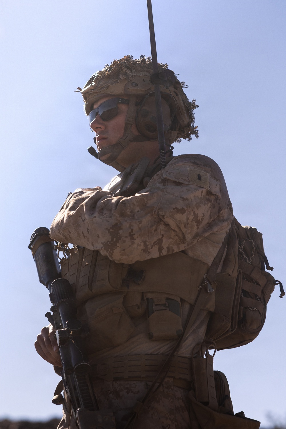 A heavily equipped military service member is photographed from a low angle against a clear blue sky. The soldier is wearing desert camouflage (MARPAT pattern consistent with USMC), full tactical gear, and is carrying a rifle. The image is dramatically lit with strong backlighting, emphasizing the silhouette and equipment of the subject.