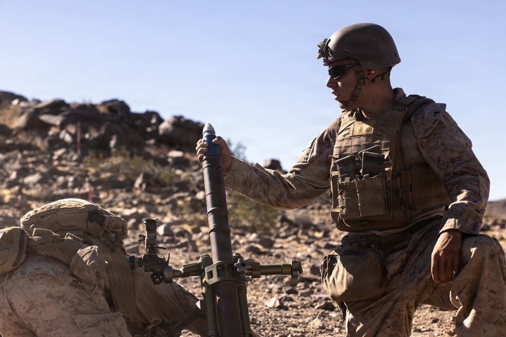 A military service member in desert camouflage uniform and tactical gear is operating a mortar system in an arid, rocky terrain. The soldier is holding a mortar round above the tube, appearing to prepare for loading. The setting and equipment suggest a training exercise or field operation in a desert environment.
