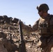 A military service member in desert camouflage uniform and tactical gear is operating a mortar system in an arid, rocky terrain. The soldier is holding a mortar round above the tube, appearing to prepare for loading. The setting and equipment suggest a training exercise or field operation in a desert environment.