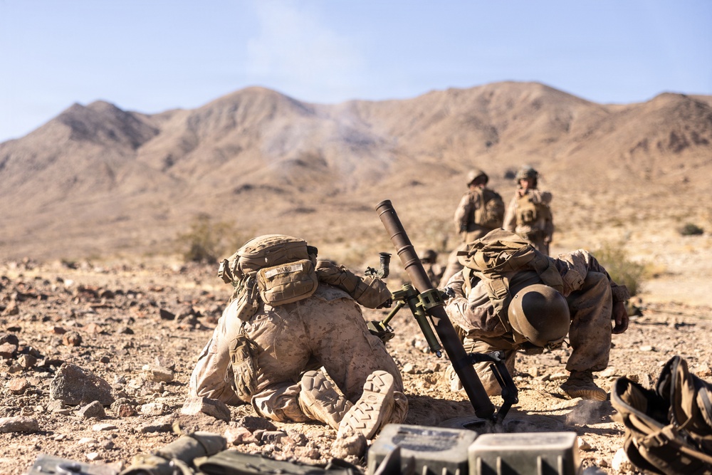 Military personnel in desert camouflage uniforms are operating a mortar system in an arid, rocky desert environment with mountains in the background. Two soldiers in the foreground are positioned around the mortar tube, while two additional personnel stand in the background. A faint smoke trail is visible above the mountains, suggesting a round has recently been fired.