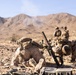 Military personnel in desert camouflage uniforms are operating a mortar system in an arid, rocky desert environment with mountains in the background. Two soldiers in the foreground are positioned around the mortar tube, while two additional personnel stand in the background. A faint smoke trail is visible above the mountains, suggesting a round has recently been fired.