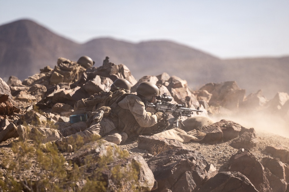 The image shows multiple soldiers in desert camouflage uniforms taking up prone firing positions behind a rocky outcropping in an arid, mountainous environment. At least two soldiers are clearly visible aiming rifles equipped with optical scopes, while additional personnel are visible in the background. The scene appears to be a military training exercise or combat operation in a desert terrain.