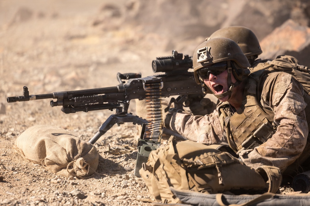 A soldier in full combat gear is positioned prone on arid, rocky terrain operating a belt-fed machine gun mounted on a bipod. The soldier appears to be shouting, possibly communicating with a second soldier visible in the background. The setting suggests a desert environment, consistent with a training exercise or combat operation.