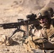 A soldier in full combat gear is positioned prone on arid, rocky terrain operating a belt-fed machine gun mounted on a bipod. The soldier appears to be shouting, possibly communicating with a second soldier visible in the background. The setting suggests a desert environment, consistent with a training exercise or combat operation.