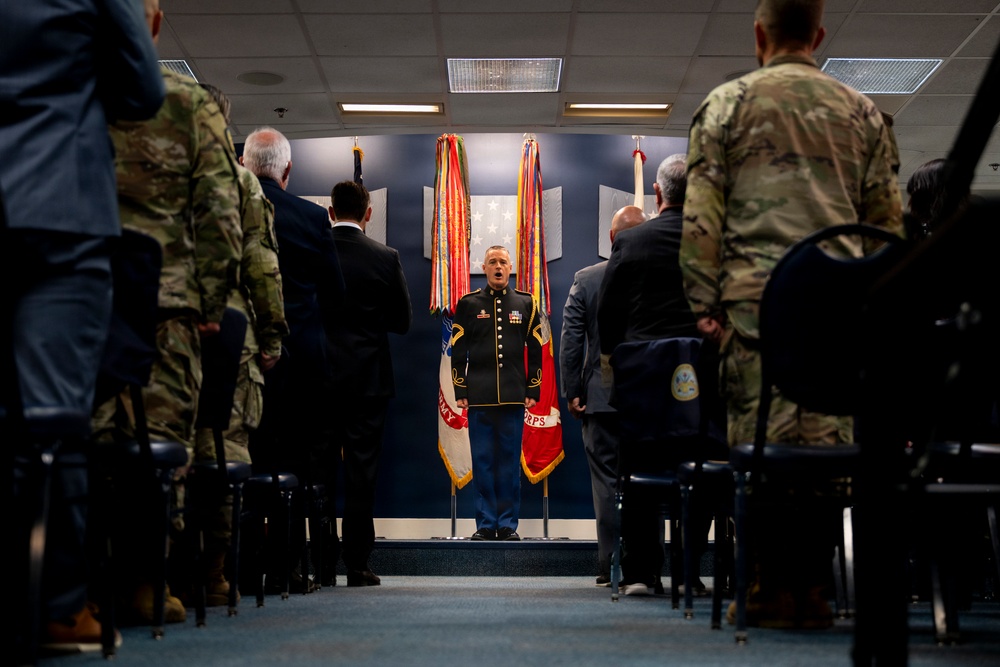 A formal military ceremony is taking place indoors, with a central figure in dress uniform standing at a podium or stage area flanked by military flags. Attendees in both military camouflage uniforms and civilian suits are standing in rows on either side of a center aisle. The setting appears to be an official military installation ceremony room with a blue-themed backdrop displaying flags and insignia.