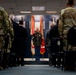 A formal military ceremony is taking place indoors, with a central figure in dress uniform standing at a podium or stage area flanked by military flags. Attendees in both military camouflage uniforms and civilian suits are standing in rows on either side of a center aisle. The setting appears to be an official military installation ceremony room with a blue-themed backdrop displaying flags and insignia.