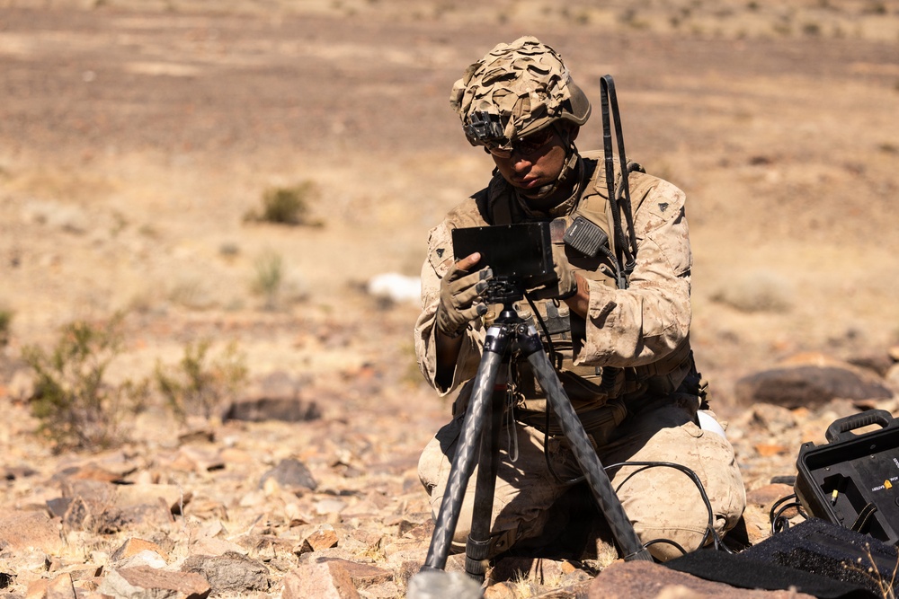 A military service member in desert camouflage uniform is kneeling in an arid, rocky terrain while operating a tablet device mounted on a tripod. The soldier is equipped with full combat gear including a helmet with camouflage netting, radio communications equipment, and tactical vest. A ruggedized equipment case is visible to the right, suggesting a field communications or surveillance operation.