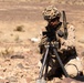 A military service member in desert camouflage uniform is kneeling in an arid, rocky terrain while operating a tablet device mounted on a tripod. The soldier is equipped with full combat gear including a helmet with camouflage netting, radio communications equipment, and tactical vest. A ruggedized equipment case is visible to the right, suggesting a field communications or surveillance operation.
