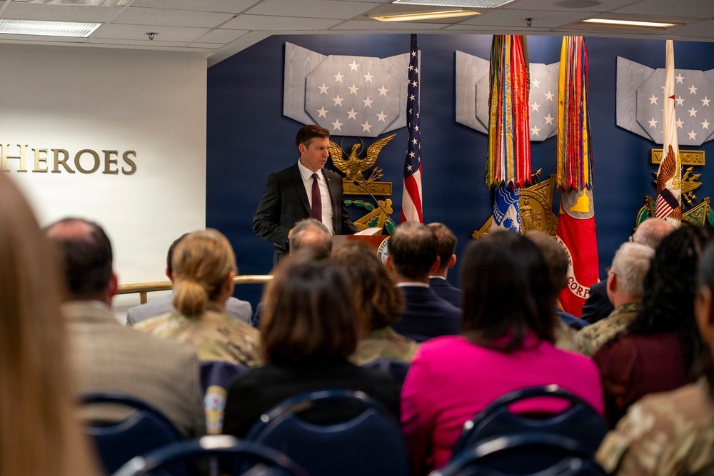 A formal military or government ceremony is taking place in an official venue, with a man in a dark suit and red tie speaking at a podium. The room features a blue wall with military insignia, multiple flags including the American flag, and an audience of mixed civilians and uniformed military personnel. The word 'HEROES' is prominently displayed in gold letters on the left wall.