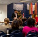 A formal military or government ceremony is taking place in an official venue, with a man in a dark suit and red tie speaking at a podium. The room features a blue wall with military insignia, multiple flags including the American flag, and an audience of mixed civilians and uniformed military personnel. The word 'HEROES' is prominently displayed in gold letters on the left wall.