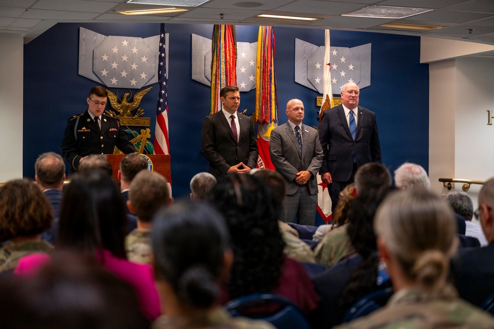 A formal military or government ceremony is taking place in an official venue with a blue-themed backdrop featuring military insignia and flags. A uniformed soldier stands at a podium while three civilians in suits stand on stage before a seated audience. The setting suggests an official recognition, award, or change-of-command type event.