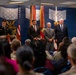 A formal military or government ceremony is taking place in an official venue with a blue-themed backdrop featuring military insignia and flags. A uniformed soldier stands at a podium while three civilians in suits stand on stage before a seated audience. The setting suggests an official recognition, award, or change-of-command type event.