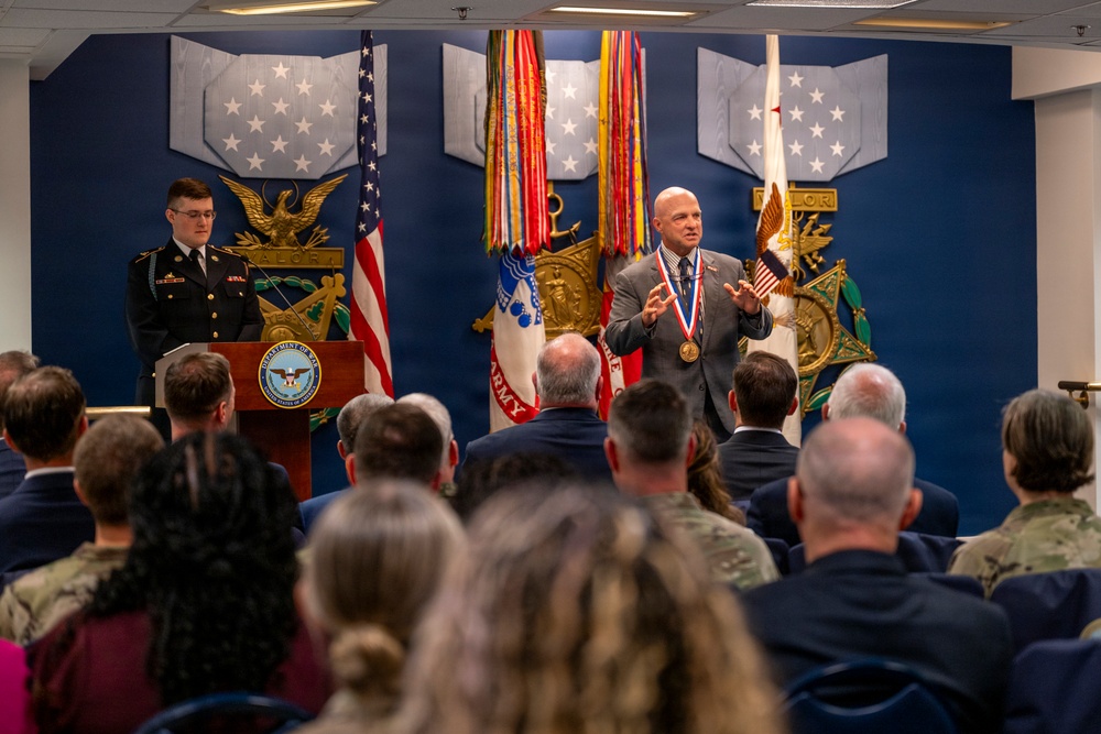 A formal military awards ceremony is taking place in what appears to be the Pentagon or a Department of Defense facility. A civilian man wearing a large medallion/medal is addressing an audience while a uniformed Army soldier stands at a podium bearing the Department of Defense seal. The backdrop features military service flags, a U.S. flag, and decorative Medal of Honor insignia on a blue wall.