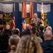 A formal military awards ceremony is taking place in what appears to be the Pentagon or a Department of Defense facility. A civilian man wearing a large medallion/medal is addressing an audience while a uniformed Army soldier stands at a podium bearing the Department of Defense seal. The backdrop features military service flags, a U.S. flag, and decorative Medal of Honor insignia on a blue wall.