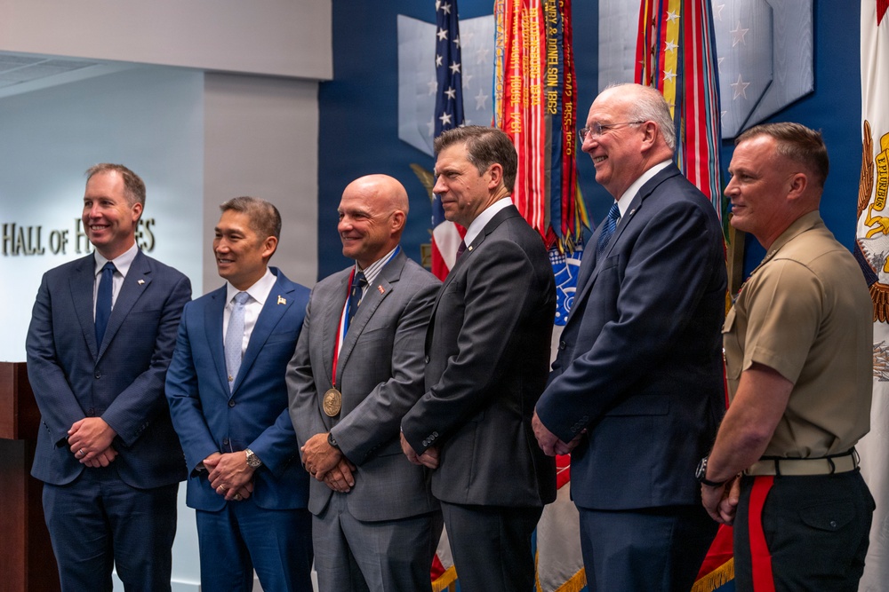 Six individuals are posed together in what appears to be a formal ceremony or award presentation, standing in front of military unit colors and flags. One individual in the center is wearing a medal with a red, white, and blue ribbon, suggesting they are the honoree. The setting includes a partial sign reading 'Hall of He[roes]' on the left wall, indicating a military or government recognition venue.