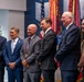 Six individuals are posed together in what appears to be a formal ceremony or award presentation, standing in front of military unit colors and flags. One individual in the center is wearing a medal with a red, white, and blue ribbon, suggesting they are the honoree. The setting includes a partial sign reading 'Hall of He[roes]' on the left wall, indicating a military or government recognition venue.