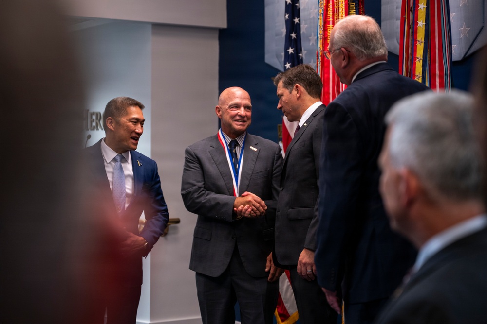 A formal ceremony appears to be taking place in what looks like a government or military institution, with multiple men in business suits gathered together. The central figure, a bald man wearing a red, white, and blue medallion/medal around his neck, is shaking hands with another suited individual. Several military or ceremonial flags are visible in the background, suggesting an official recognition or award event.