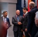 A formal ceremony appears to be taking place in what looks like a government or military institution, with multiple men in business suits gathered together. The central figure, a bald man wearing a red, white, and blue medallion/medal around his neck, is shaking hands with another suited individual. Several military or ceremonial flags are visible in the background, suggesting an official recognition or award event.