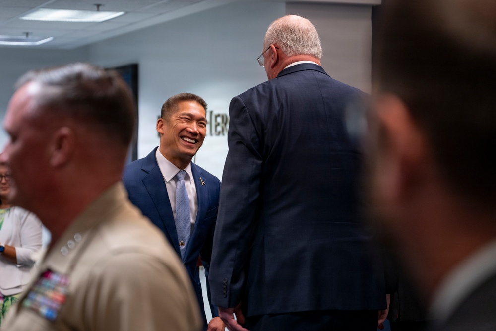 A professional gathering or meeting in an office or conference room setting. A man in a navy blue suit and light blue tie is smiling broadly while conversing with an older man whose back is turned to the camera. Military personnel in tan/khaki uniform are visible in the foreground.