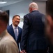 A professional gathering or meeting in an office or conference room setting. A man in a navy blue suit and light blue tie is smiling broadly while conversing with an older man whose back is turned to the camera. Military personnel in tan/khaki uniform are visible in the foreground.