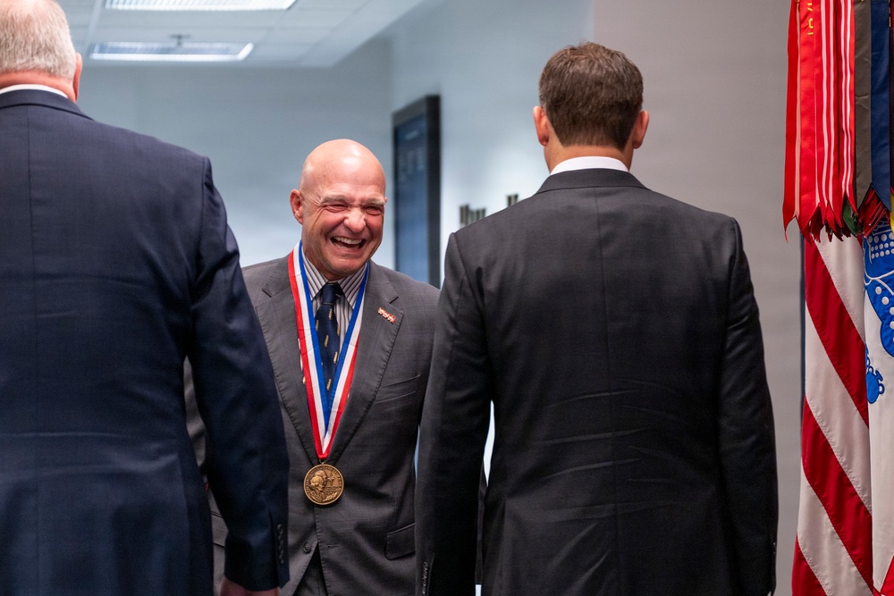 A bald man wearing a gold medal on a red, white, and blue ribbon lanyard is laughing and conversing with two other men in dark suits in what appears to be an official or ceremonial setting. The medal appears to be a distinguished honor or award. Flags, including what appears to be a U.S.-related flag, are visible in the background.